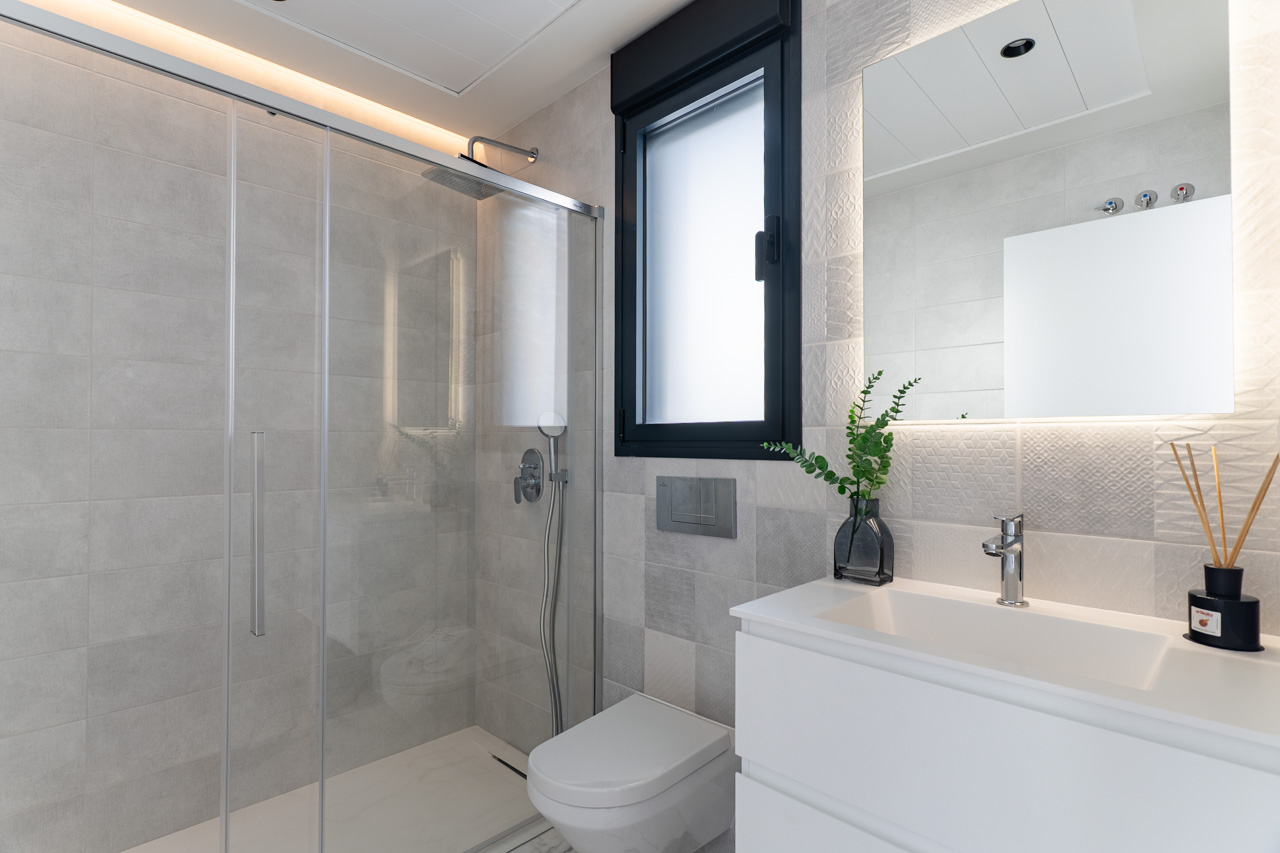 Salle de bain moderne avec douche à l'italienne, paroi en verre, carrelage texturé gris, meuble vasque blanc et miroir rétroéclairé par LED.