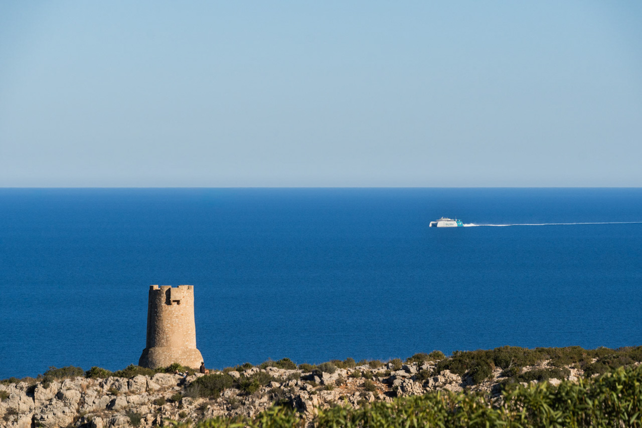 Vues panoramiques du littoral de Las Rotas à Dénia, mettant en valeur la tour historique Torre del Gerro et un ferry Balearia naviguant sur la mer Méditerranée.