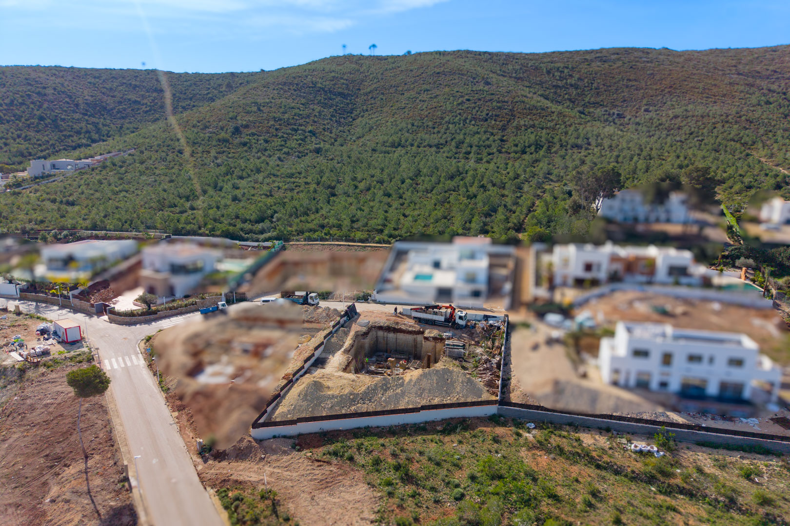 Vue aérienne de la parcelle en phase de construction, montrant l'excavation du sous-sol et les fondations au sein d'un quartier résidentiel entouré de collines verdoyantes.
