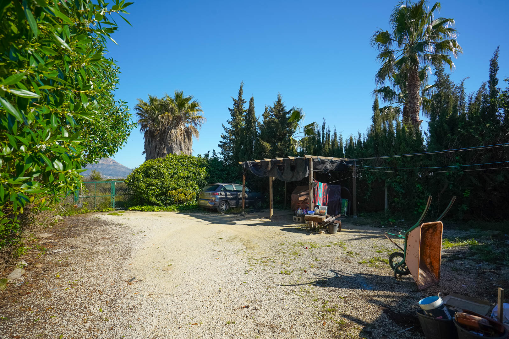 Aire de stationnement en gravier avec abri en bois, entourée de palmiers et de cyprès, offrant une vue dégagée sur le massif du Montgó.