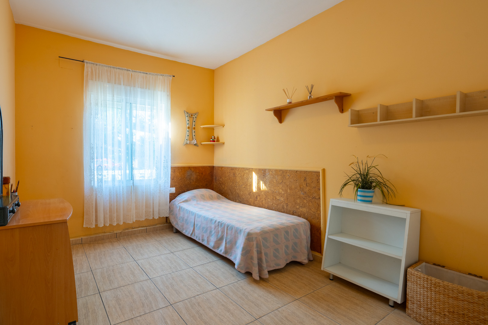 Chambre avec murs ocre, sol en grès cérame, lit simple et étagères en bois. La fenêtre est ornée de rideaux en dentelle laissant passer la lumière naturelle.