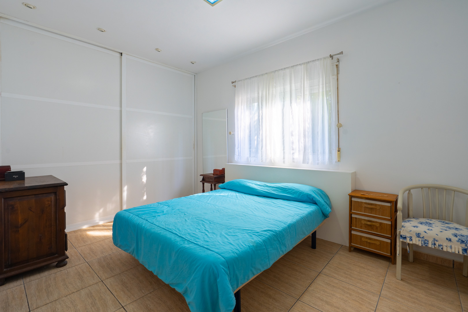 Chambre lumineuse avec de grands placards intégrés blancs, sol en carrelage, fenêtre avec voilages et mobilier en bois de style classique.