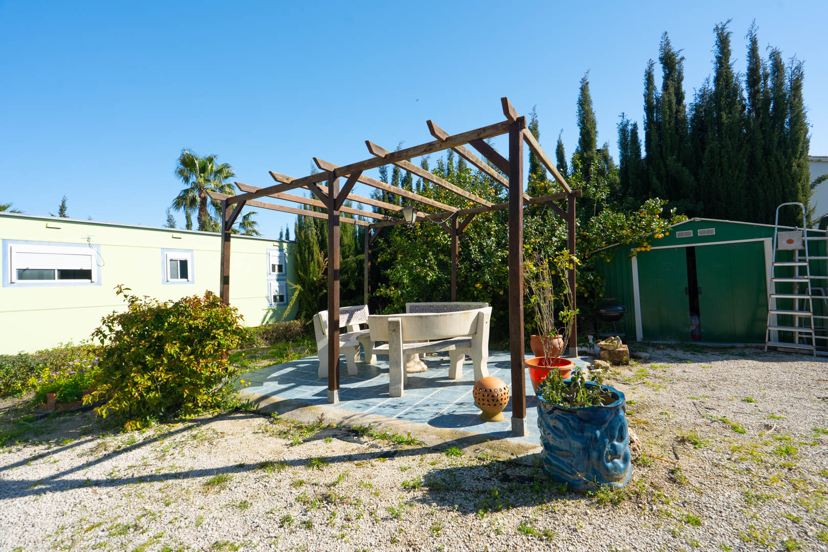 Coin salon extérieur avec pergola en bois et bancs en pierre sur terrasse carrelée, situé entre l'abri de jardin et la maison d'invités préfabriquée.