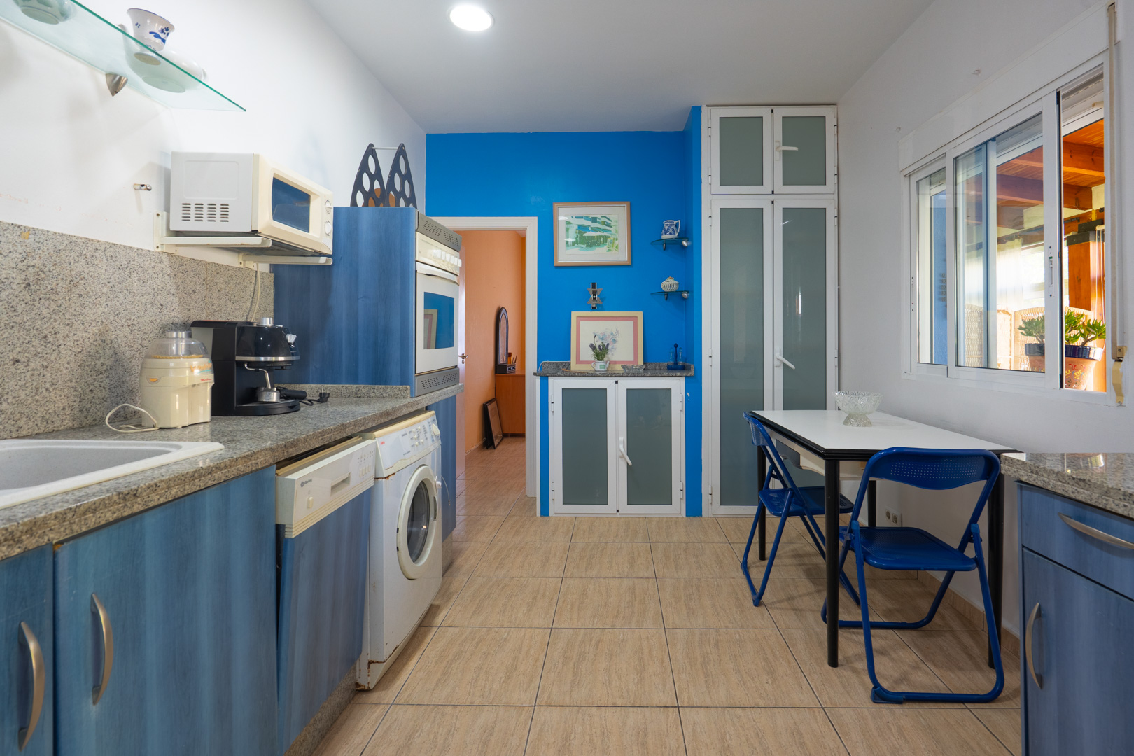 Cuisine équipée avec des placards bleus, des plans de travail en granit gris, un coin repas et des appareils intégrés sous un éclairage encastré.