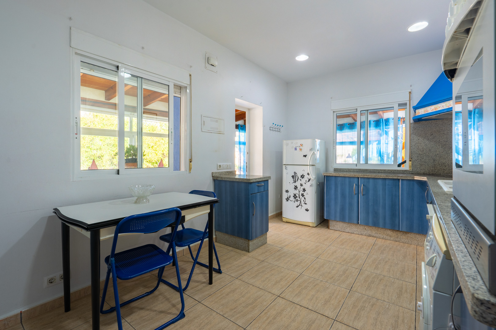 Cuisine fonctionnelle avec des meubles bleus, des plans de travail en granit et un coin repas. Elle dispose de spots encastrés et de fenêtres donnant sur l'extérieur.