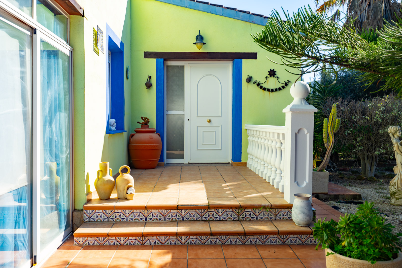 Entrée principale avec porte blanche, cadres bleus et contremarches en carrelage décoratif. La terrasse en terre cuite dispose d'une balustrade blanche et de poteries.