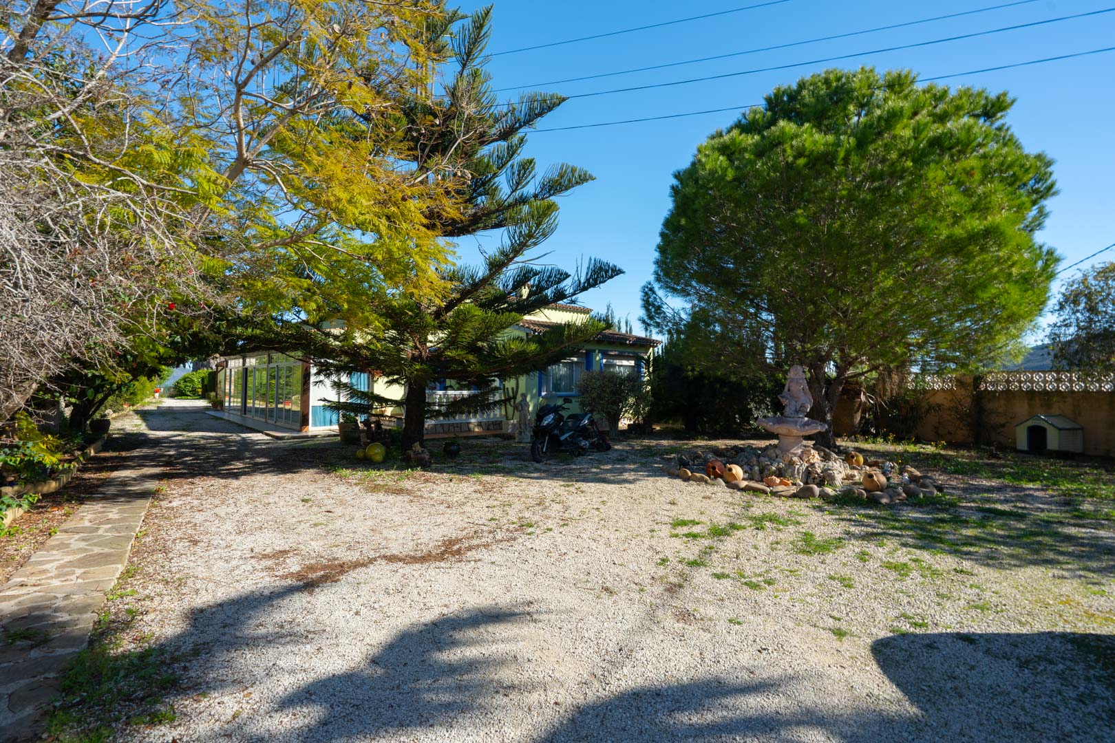 Espace d'entrée en gravier avec des pins matures et une fontaine décorative, offrant une vue sur la villa et sa naya fermée par des vitrages.