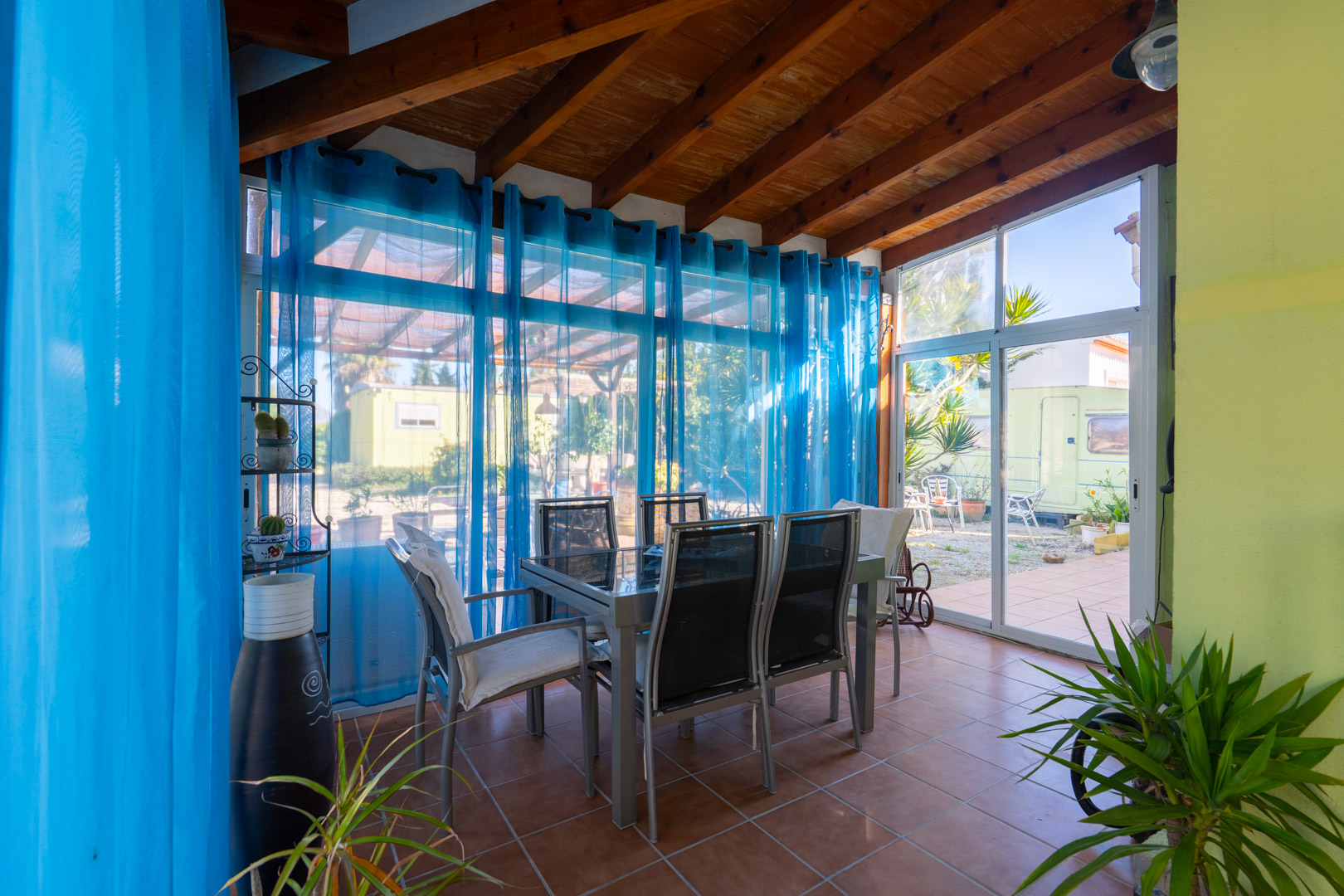 Naya vitrée avec poutres en bois apparentes, sol en terre cuite et espace salle à manger offrant un accès direct au jardin.