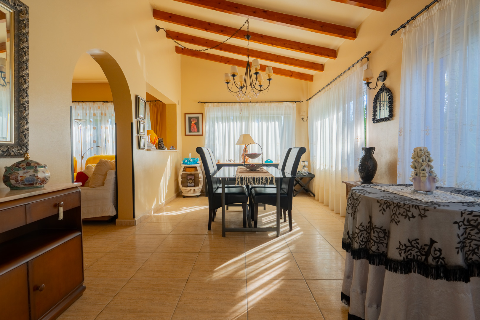 Salle à manger avec plafonds à poutres apparentes, sol en grès cérame et une arche traditionnelle menant au salon baigné de lumière naturelle.