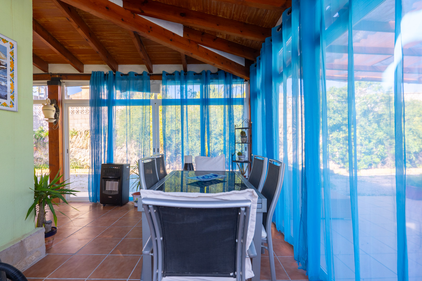 Salle à manger sous naya vitrée avec poutres en bois apparentes, sol en terre cuite et rideaux bleus offrant une vue sur l'extérieur.