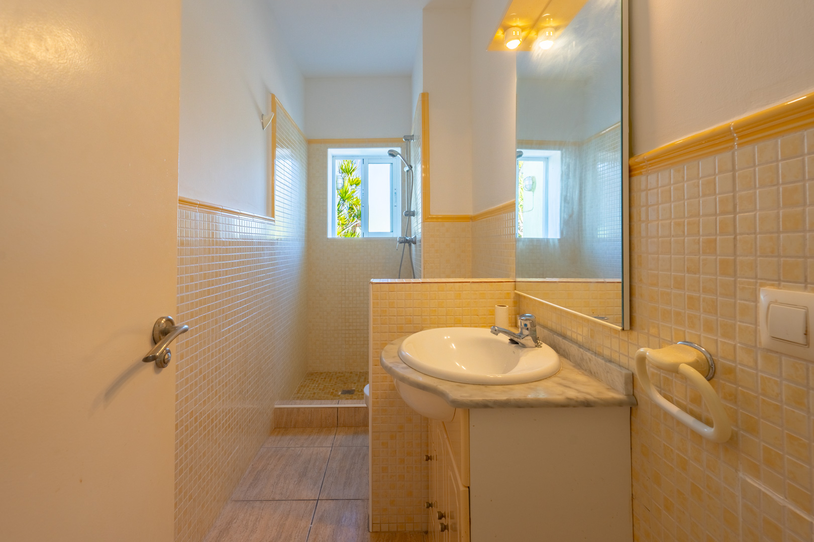 Salle de bain avec douche à l'italienne, carrelage en mosaïque jaune et crème, meuble vasque avec plan en marbre et éclairage intégré.