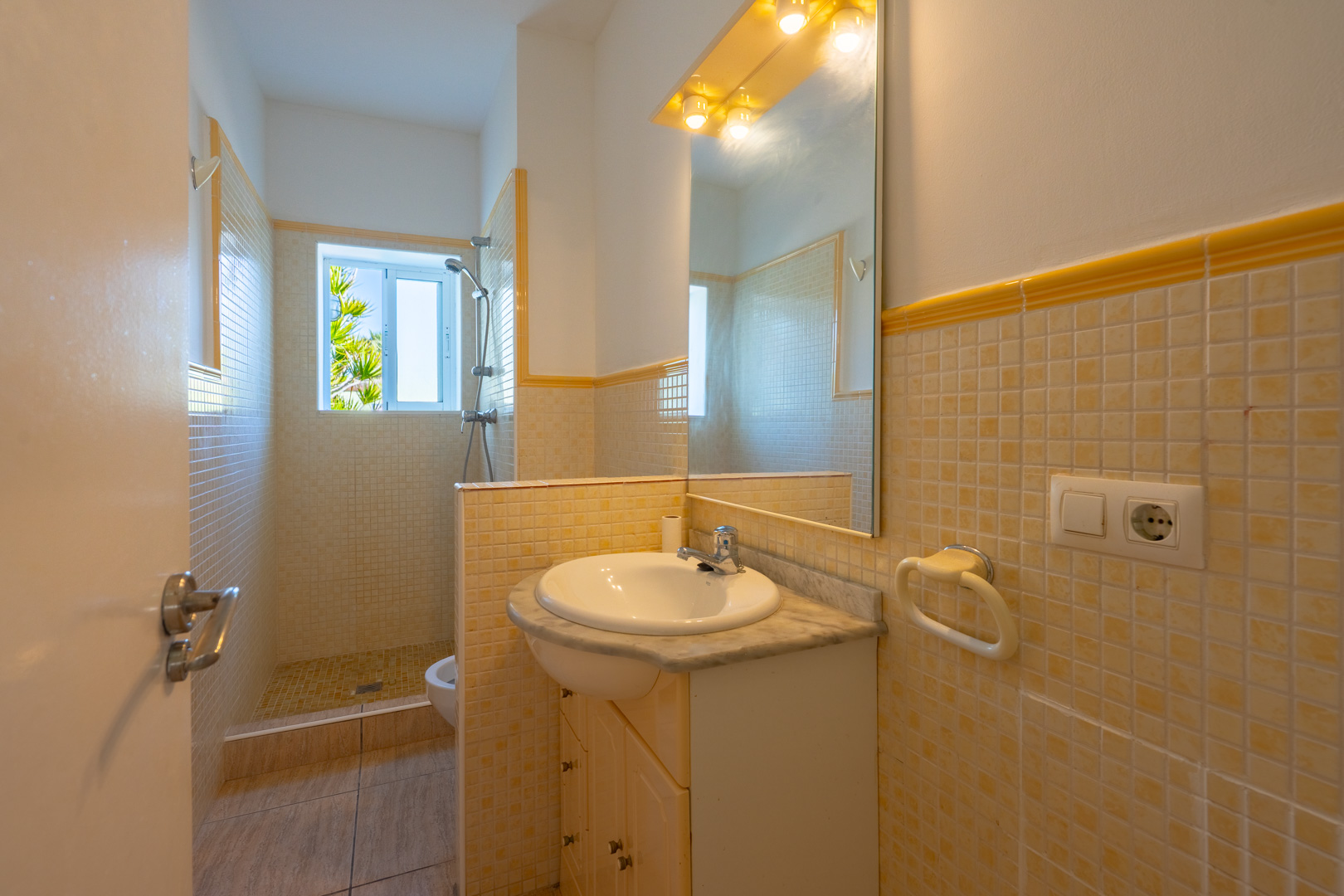 Salle de bains avec douche à l'italienne en mosaïque, meuble vasque en marbre et grand miroir éclairé. Une fenêtre offre une lumière naturelle.