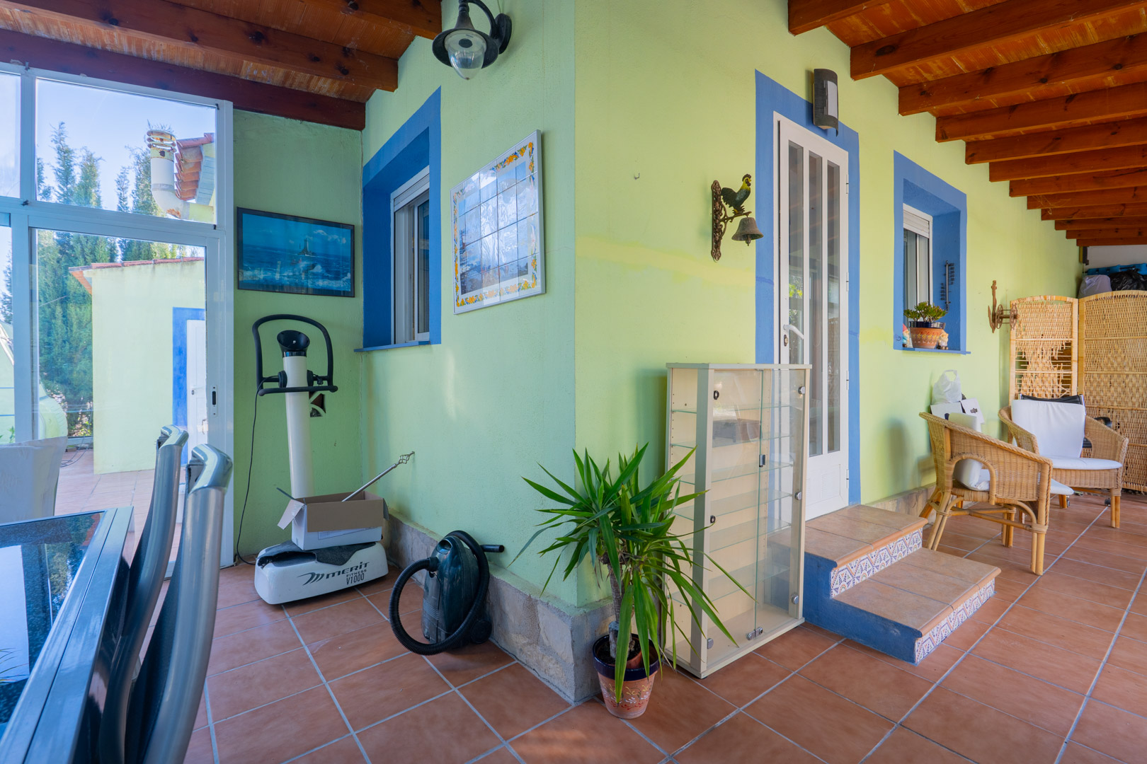 Terrasse couverte avec poutres en bois apparentes, murs colorés vert et bleu, sol en terre cuite et mobilier en osier dans un style traditionnel.