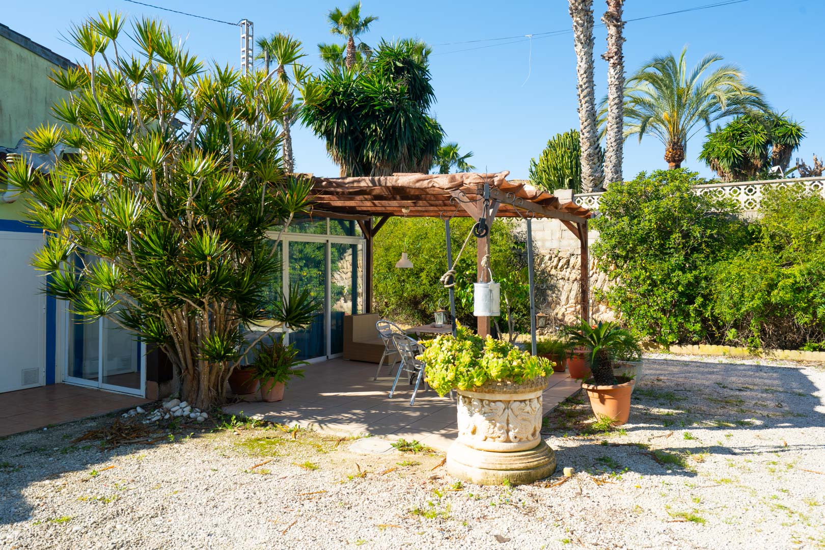 Terrasse extérieure avec pergola en bois, coin repas et végétation méditerranéenne luxuriante, agrémentée de jardinières en pierre sculptée.