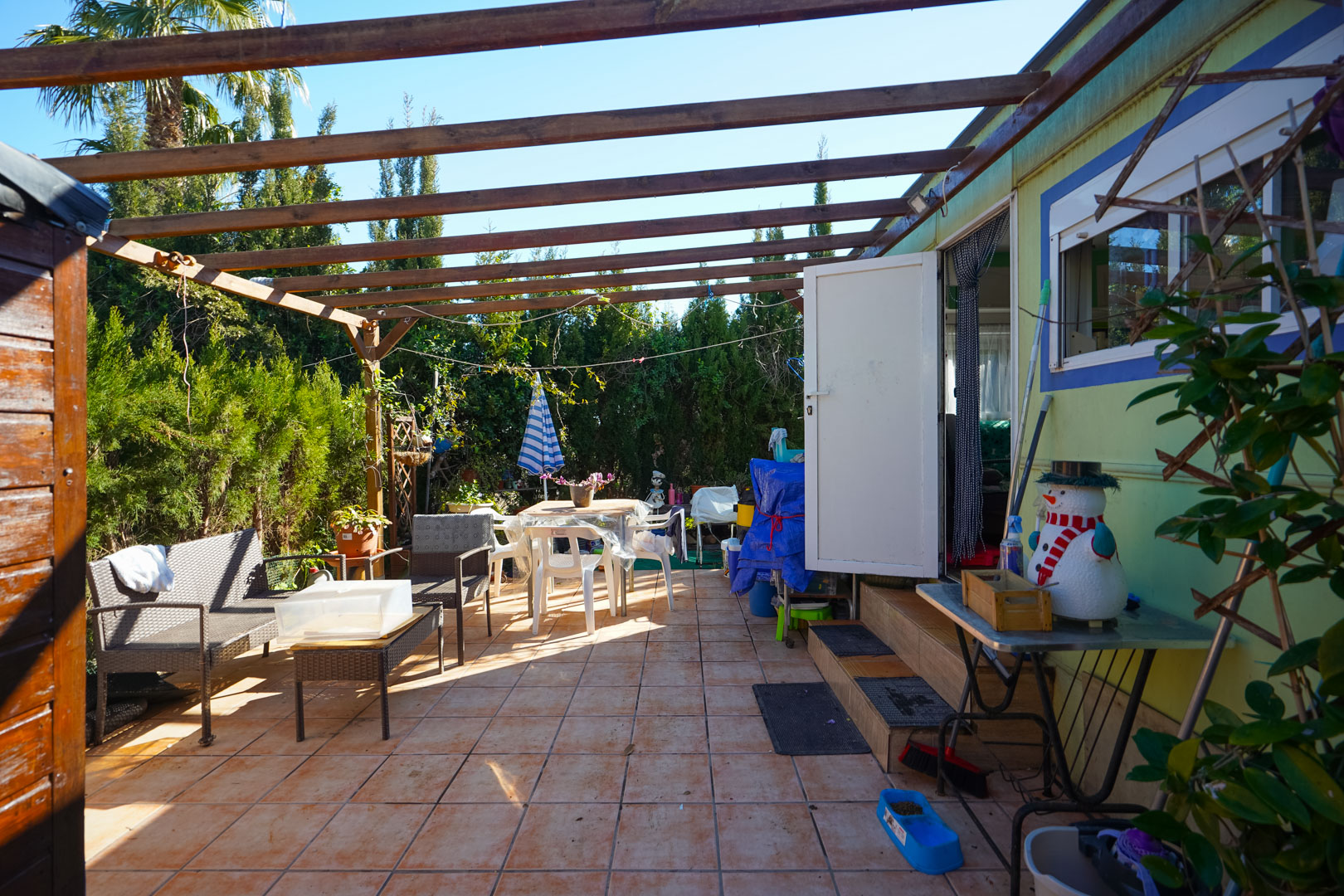 Terrasse extérieure de l'habitation préfabriquée avec sol en céramique, pergola à poutres en bois et coin salon entouré d'une haie dense.