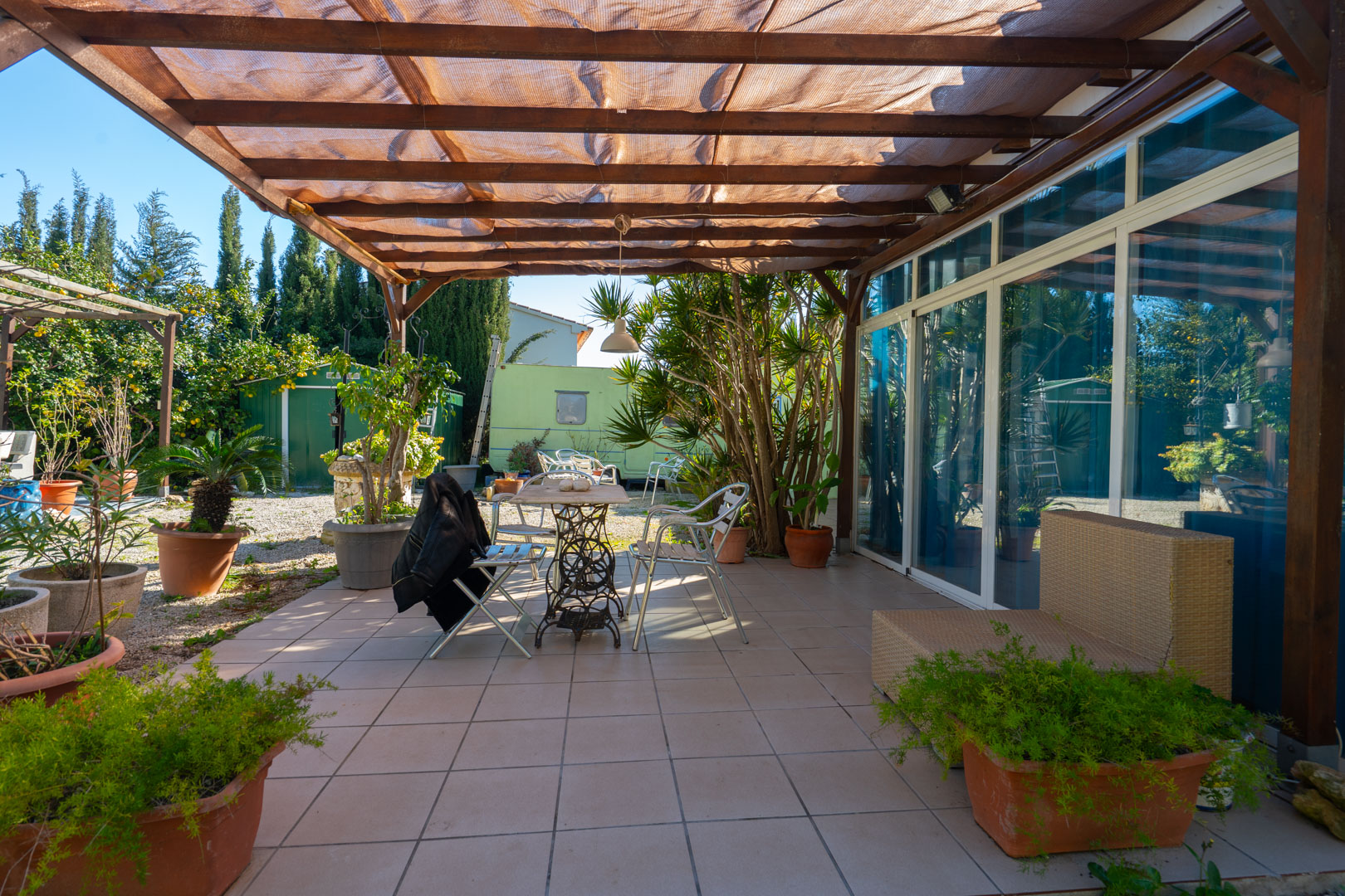 Terrasse pavée avec pergola en bois et voile d'ombrage, équipée de mobilier d'extérieur et bordée de plantes en pots face au jardin.