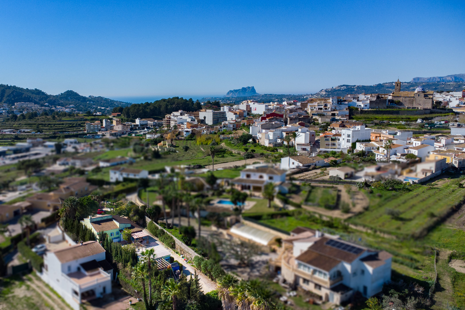 Vue aérienne panoramique de Benitachell avec le Peñón de Ifach à l'horizon. On aperçoit le centre-ville, l'église paroissiale et le paysage rural bordant la mer Méditerranée.