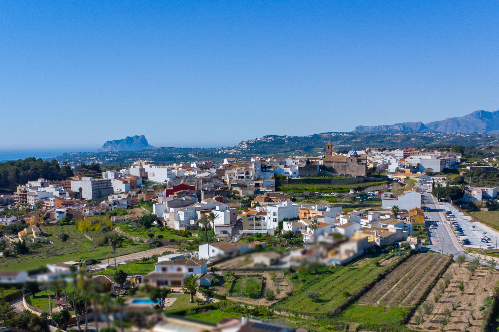 Vue aérienne panoramique de Benitachell illustrant le centre historique, l'église et le Peñón de Ifach bordant la mer Méditerranée.