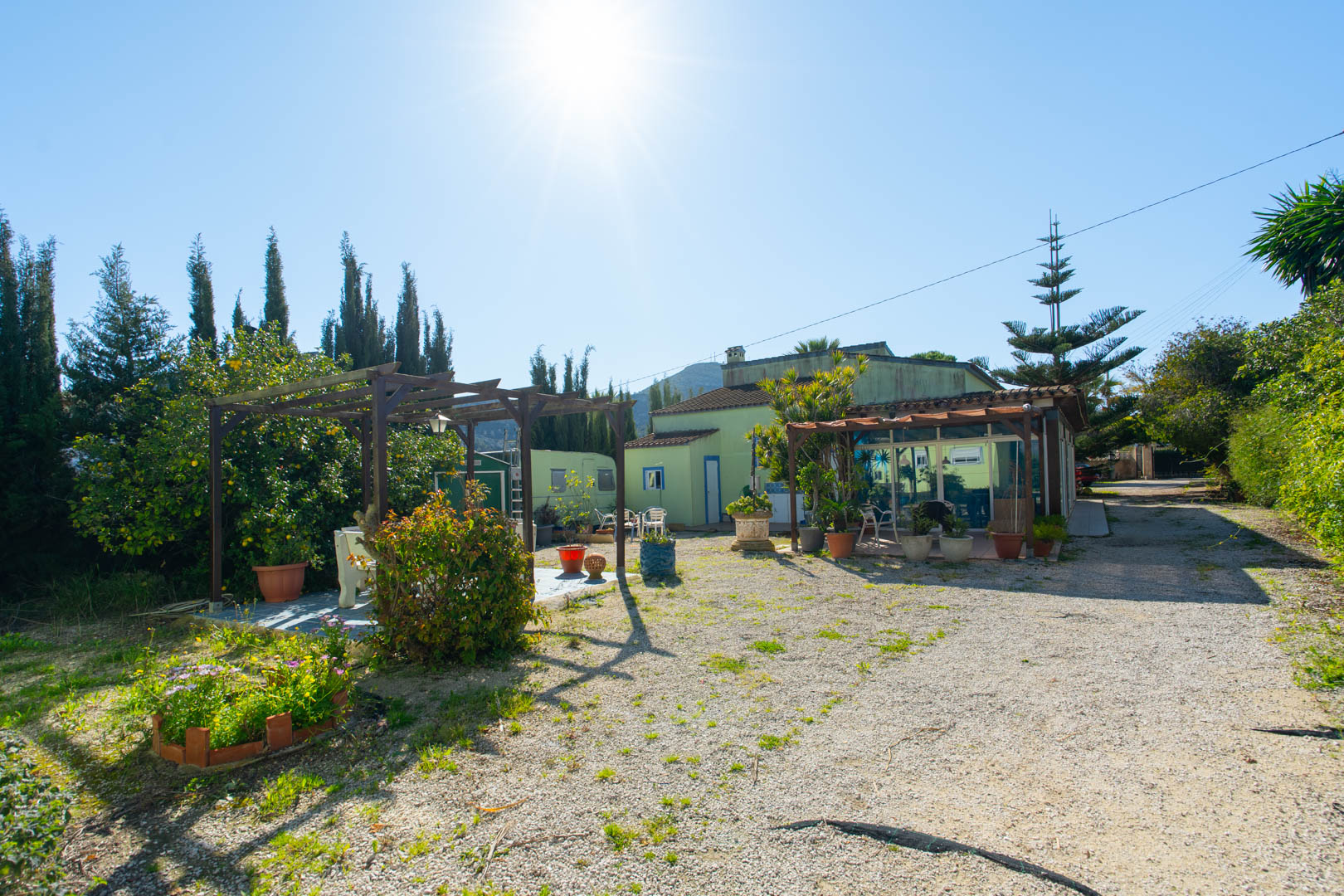 Vue extérieure du jardin rustique avec une pergola en bois, un coin salon et la façade de la maison principale dotée d'une naya vitrée.