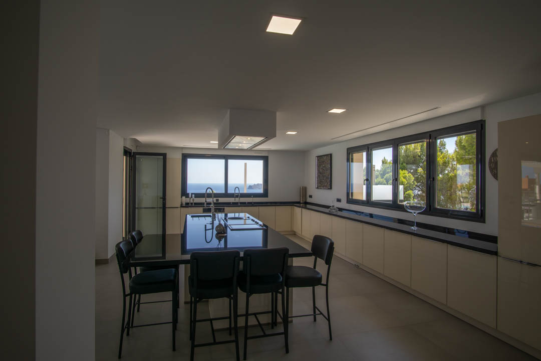 Cuisine moderne avec îlot central en granit noir, armoires minimalistes crème et larges fenêtres offrant une vue panoramique sur la mer Méditerranée.