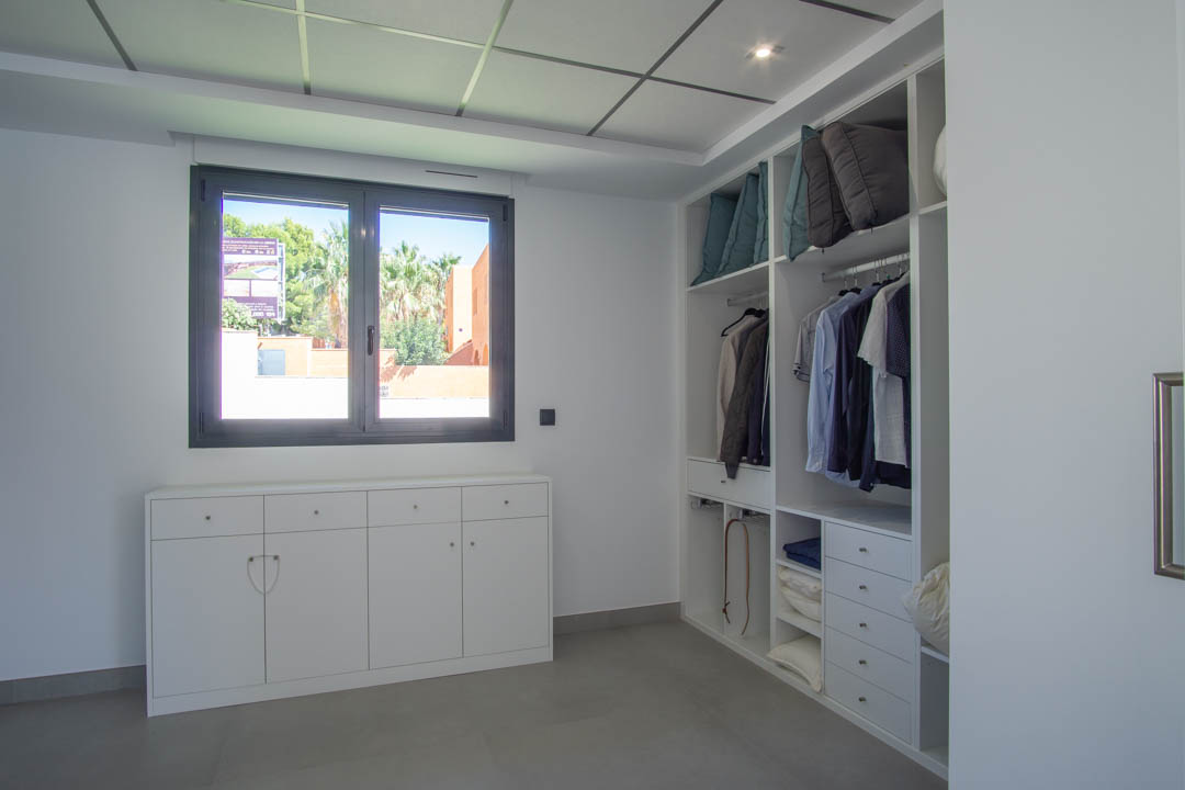 Dressing avec rangements sur mesure en blanc, tiroirs intégrés et sol en grès gris. Une fenêtre à cadre anthracite offre une vue dégagée et une luminosité naturelle.