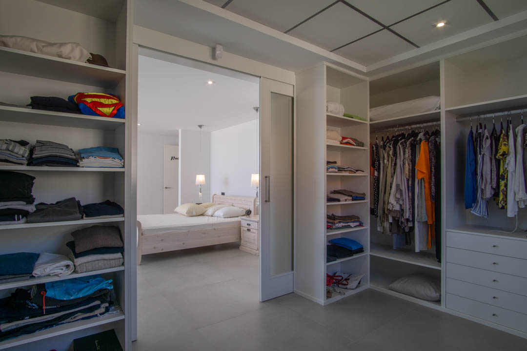 Dressing de 12m2 avec étagères blanches sur mesure, tiroirs intégrés et porte coulissante vitrée menant à la chambre principale avec éclairage encastré.