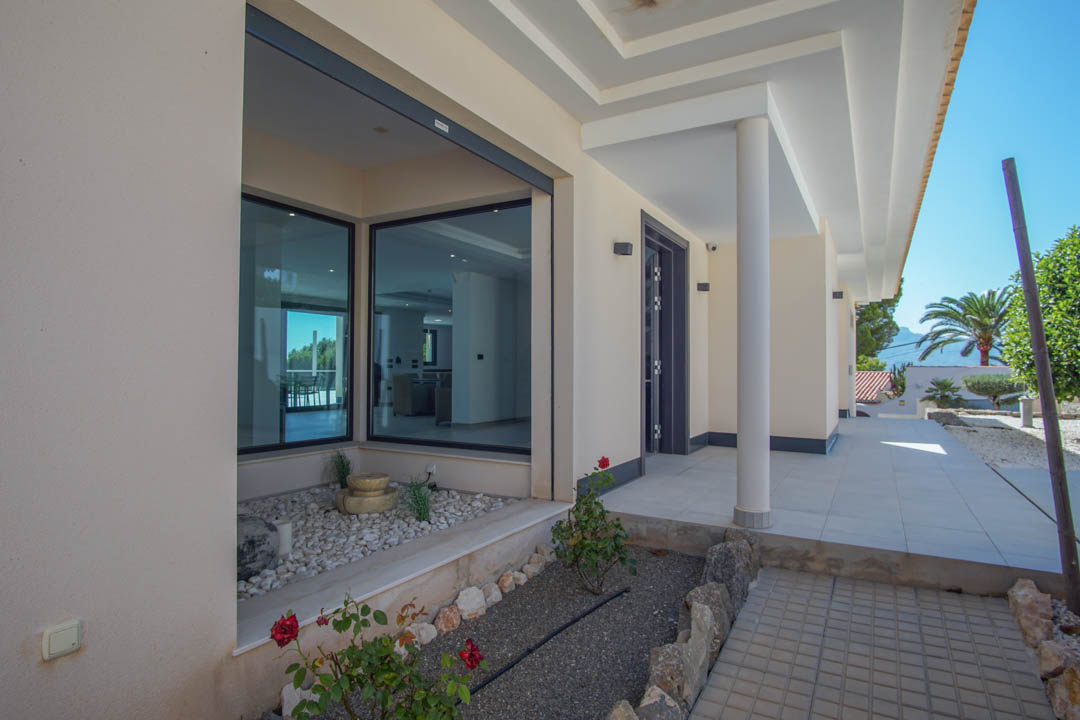 Entrée principale avec porche couvert, colonnes blanches et grandes baies vitrées. Un jardin zen avec galets blancs et fontaine orne l'accès à cette villa moderne.