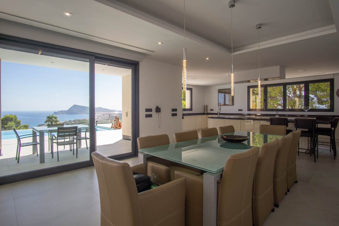 Espace dînatoire ouvert avec table en verre dépoli, cuisine contemporaine et accès à la terrasse avec vue panoramique sur la côte et la piscine.
