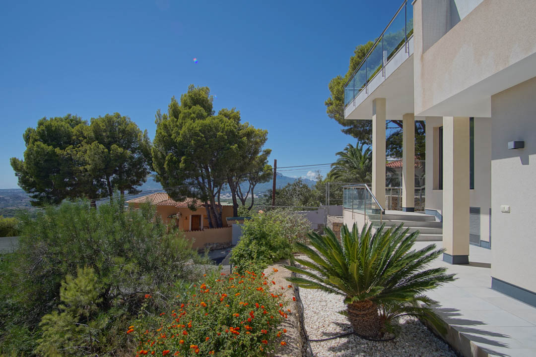 Extérieur de la villa avec colonnes blanches, balustrades en verre et jardin paysager comprenant des cycas et des arbustes à fleurs en bordure de terrasse.