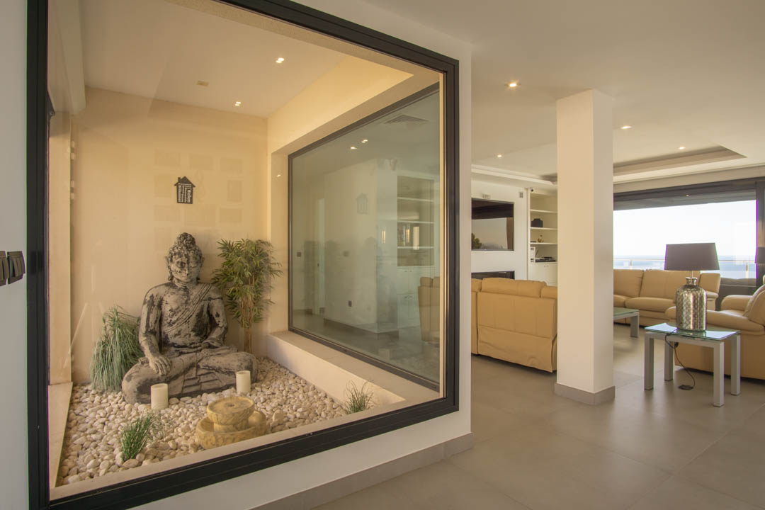 Jardin intérieur zen vitré avec statue de Bouddha et galets blancs, ouvert sur un vaste salon aux tons neutres avec éclairage encastré au plafond.