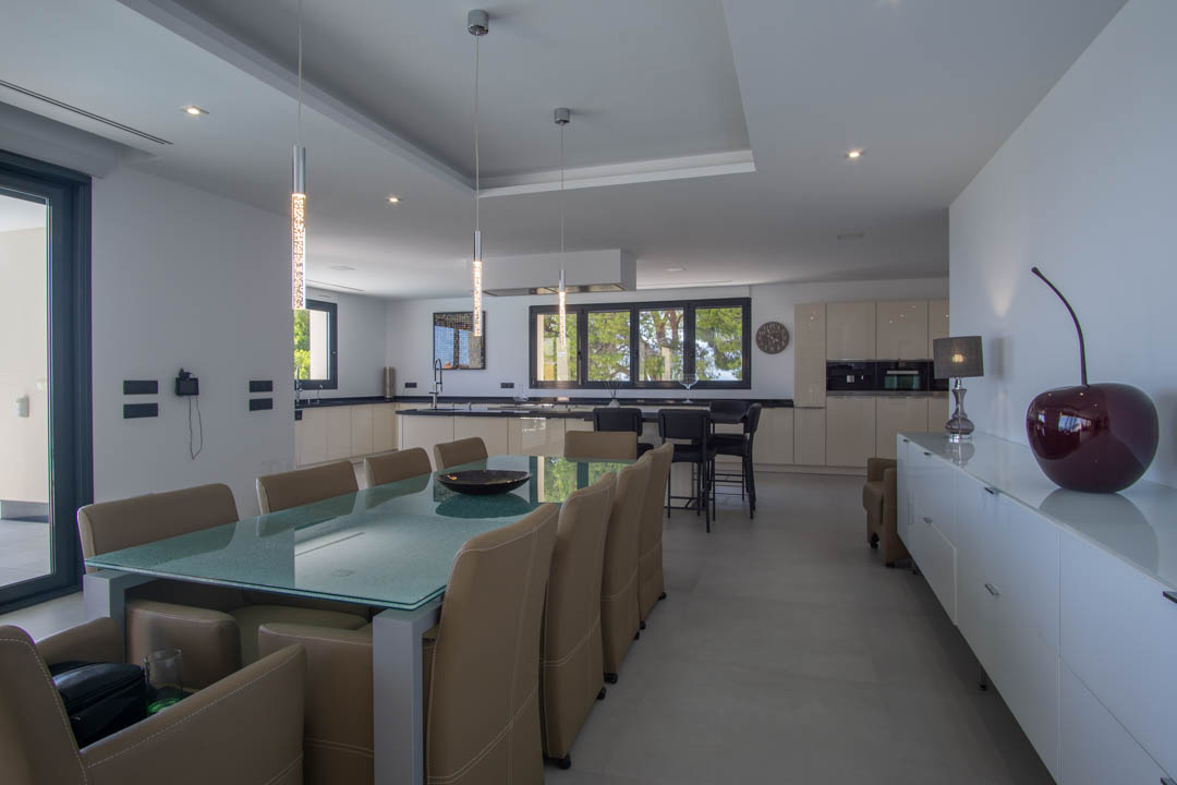 Salle à manger à aire ouverte avec table en verre et chaises en cuir, intégrée à une cuisine moderne équipée de plans de travail sombres et d'un éclairage encastré.