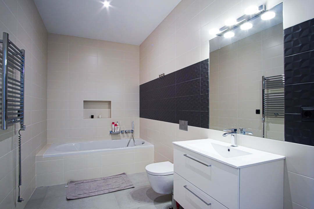 Salle de bain moderne avec baignoire encastrée, meuble vasque blanc, WC suspendu et carrelage beige agrémenté d'une bande texturée gris foncé.