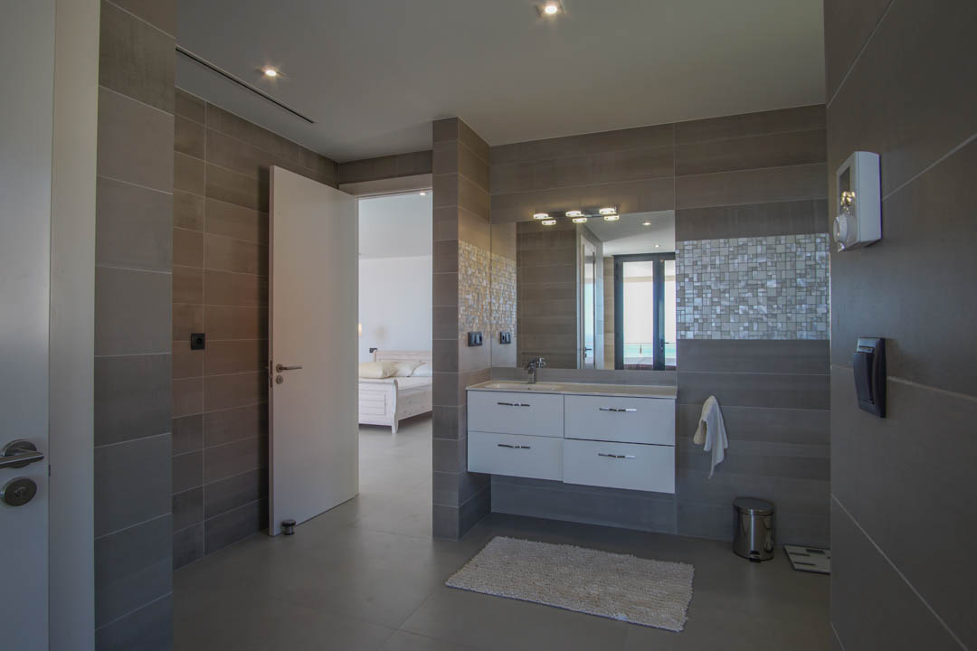 Salle de bains contemporaine avec carrelage gris, meuble vasque suspendu et mosaïque. La pièce s'ouvre sur la chambre principale offrant une perspective vers la mer.