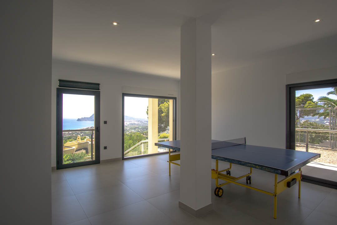 Salle de jeux avec table de ping-pong, carrelage gris et grandes baies vitrées offrant une vue panoramique sur la mer Méditerranée et la côte d'Altea.
