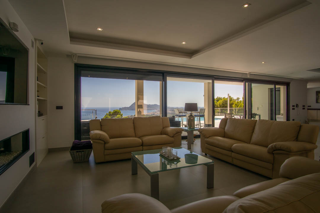 Salon spacieux avec canapés en cuir beige et grandes baies vitrées coulissantes offrant une vue panoramique sur la baie d'Altea. Comprend une cheminée intégrée et un éclairage encastré.