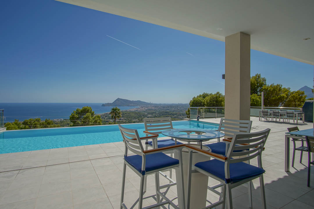 Terrasse spacieuse avec piscine à débordement offrant une vue panoramique sur la mer. Comprend un coin salon moderne avec mobilier blanc et bleu.