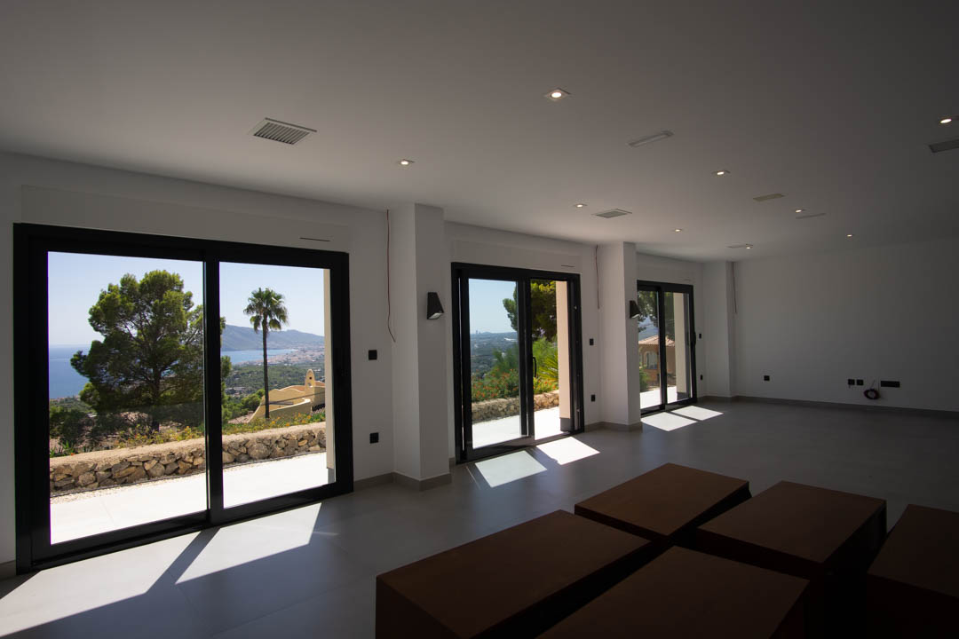 Vaste espace de vie ouvert avec de grandes baies vitrées coulissantes à cadre noir, éclairage encastré au plafond et vue panoramique sur la mer et le littoral d'Altea.