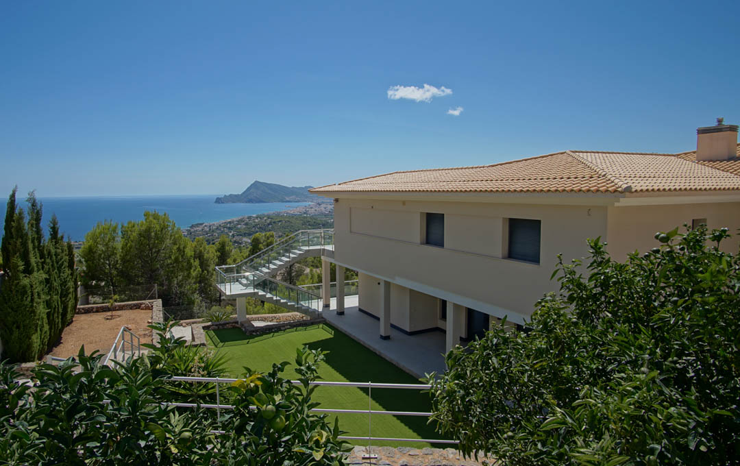 Vue extérieure de la villa avec jardin en gazon synthétique, agrumes et escalier en verre, offrant un panorama sur la baie d'Altea et la mer.