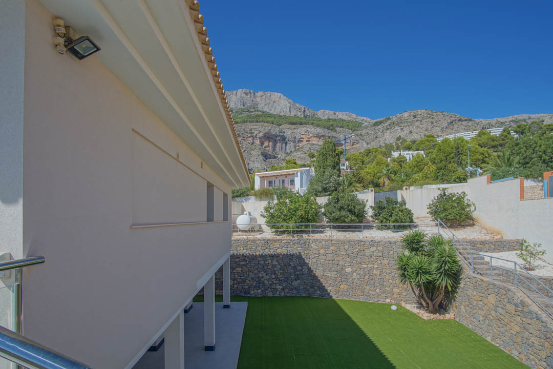Vue extérieure du jardin inférieur avec gazon synthétique, murs en pierre naturelle et escaliers métalliques, avec la Sierra de Altea en arrière-plan.