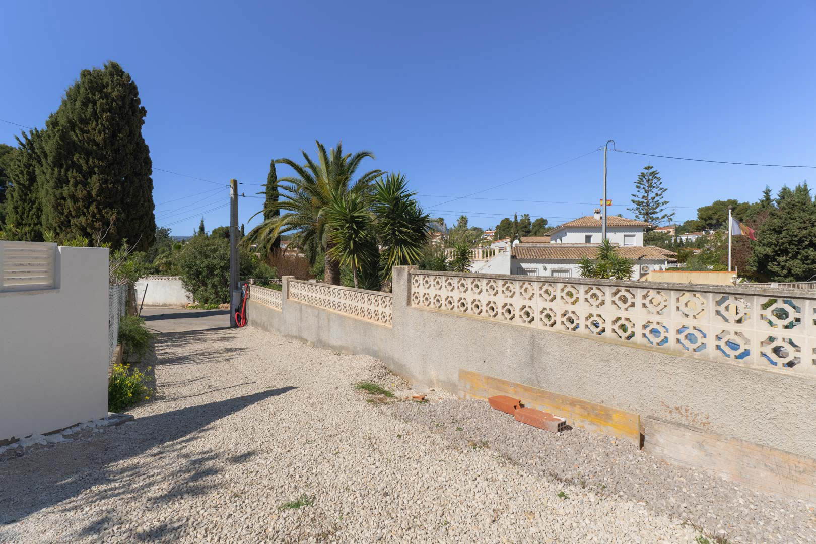 Accès extérieur à la propriété avec sol en gravier blanc et mur de clôture en blocs décoratifs, entouré de palmiers et de végétation méditerranéenne.