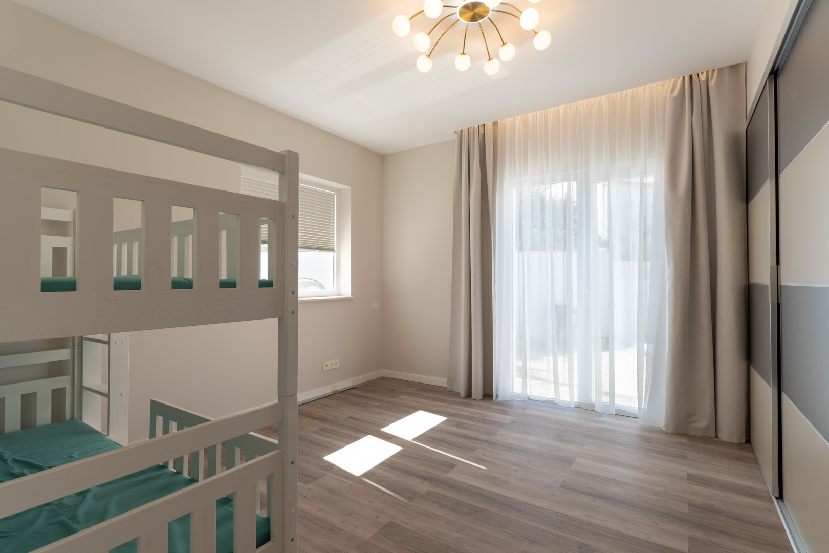 Chambre avec lits superposés en bois blanc, sol stratifié et grand placard intégré. La lumière naturelle pénètre par de larges fenêtres avec voilages.