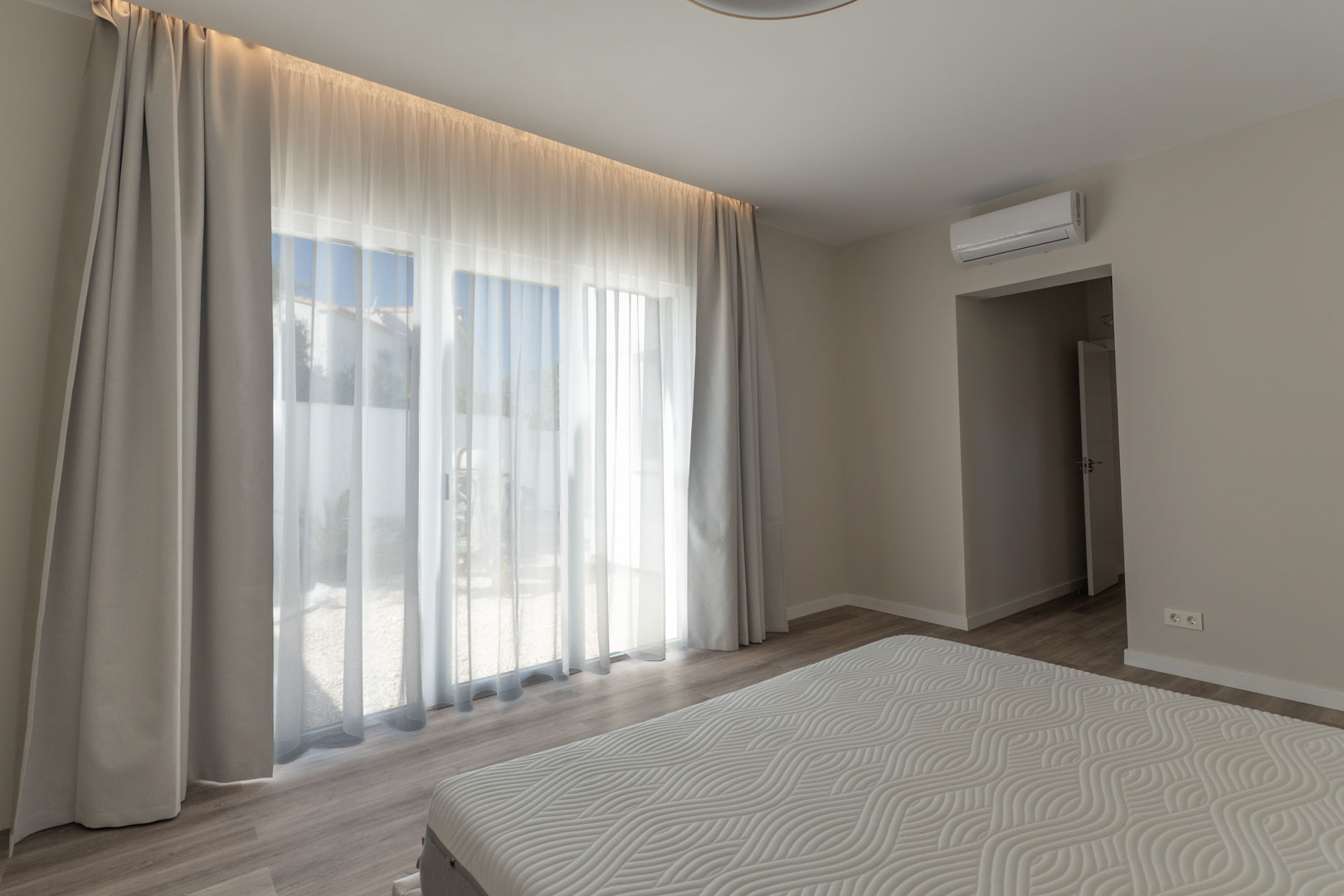 Chambre minimaliste avec de grandes baies vitrées, éclairage LED intégré et climatisation. Elle dispose d'un sol en parquet clair et d'un accès direct vers l'extérieur.