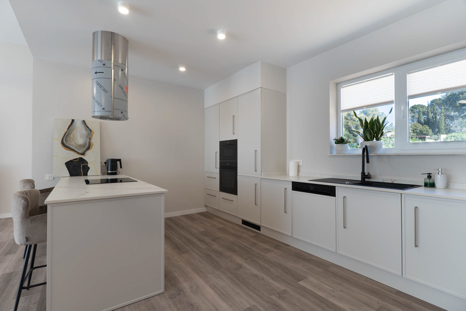 Cuisine moderne avec îlot central, plaque à induction et hotte cylindrique. Les meubles blancs contrastent avec l'évier noir et le sol en bois stratifié chaleureux.