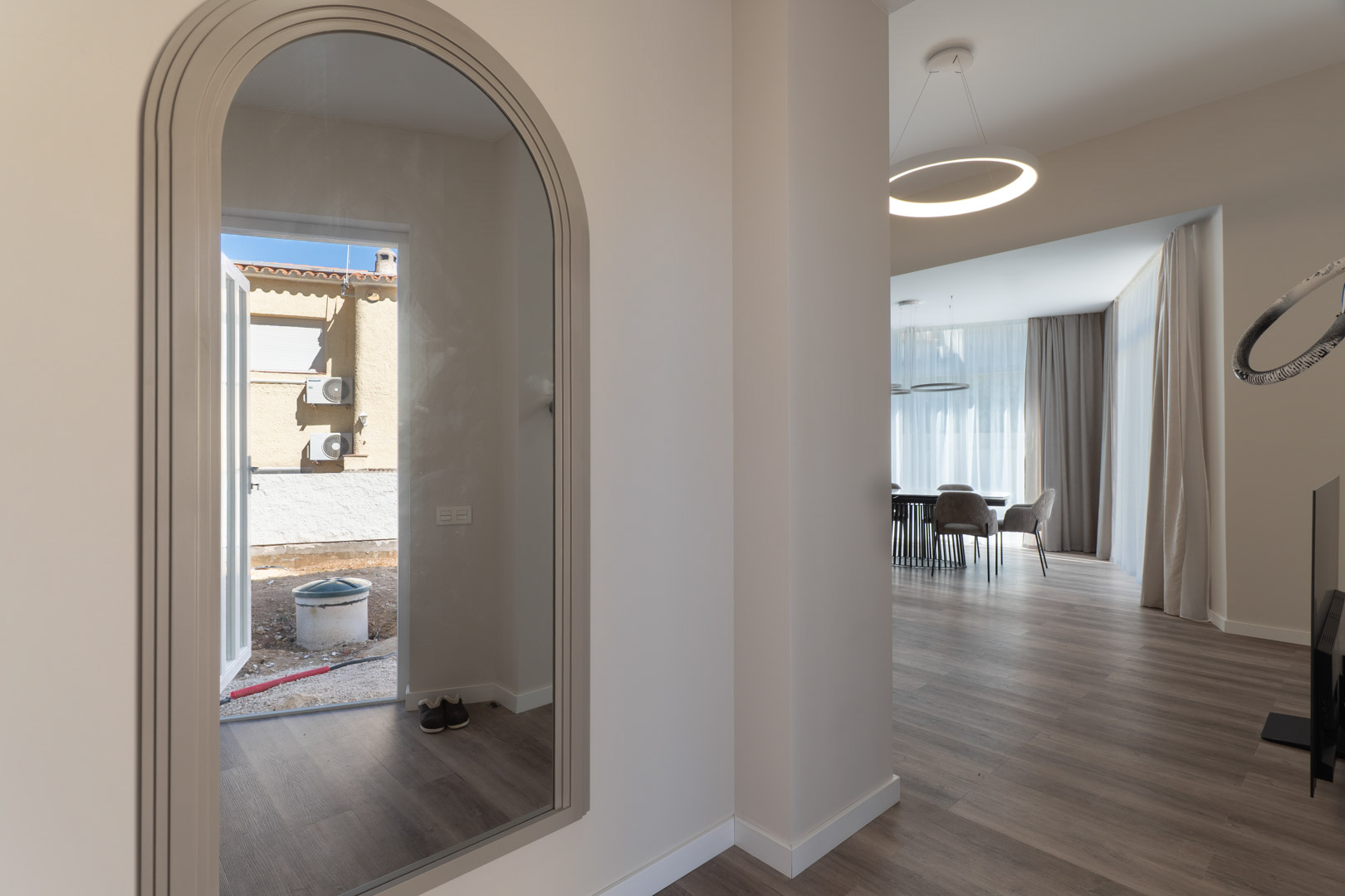 Entrée avec miroir cintré reflétant l'extérieur, s'ouvrant sur un espace de vie minimaliste avec parquet clair et suspension LED circulaire.