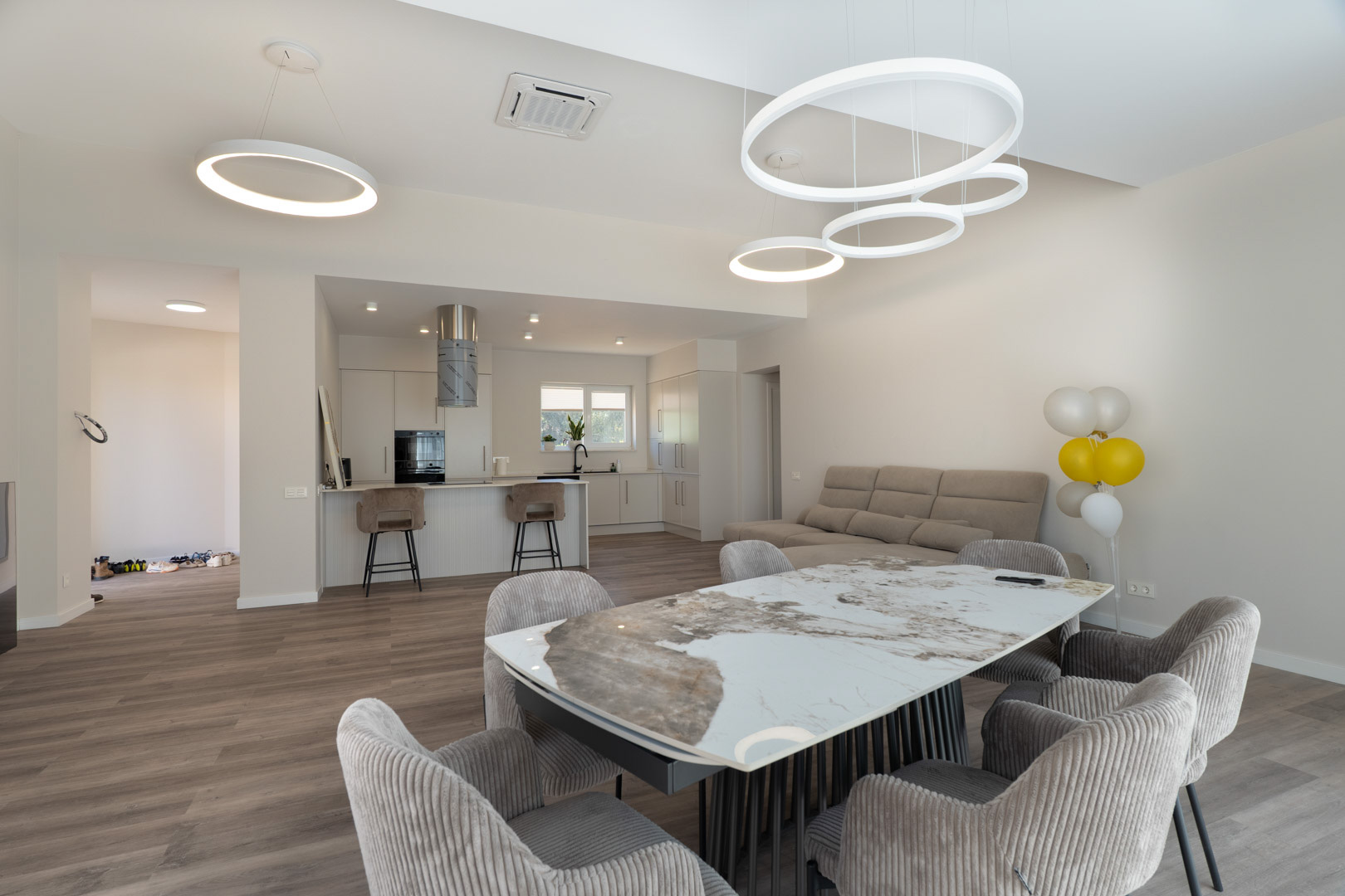 Espace de vie ouvert comprenant une table à manger en marbre, des chaises rembourrées et une cuisine blanche minimaliste avec îlot et luminaires LED circulaires.