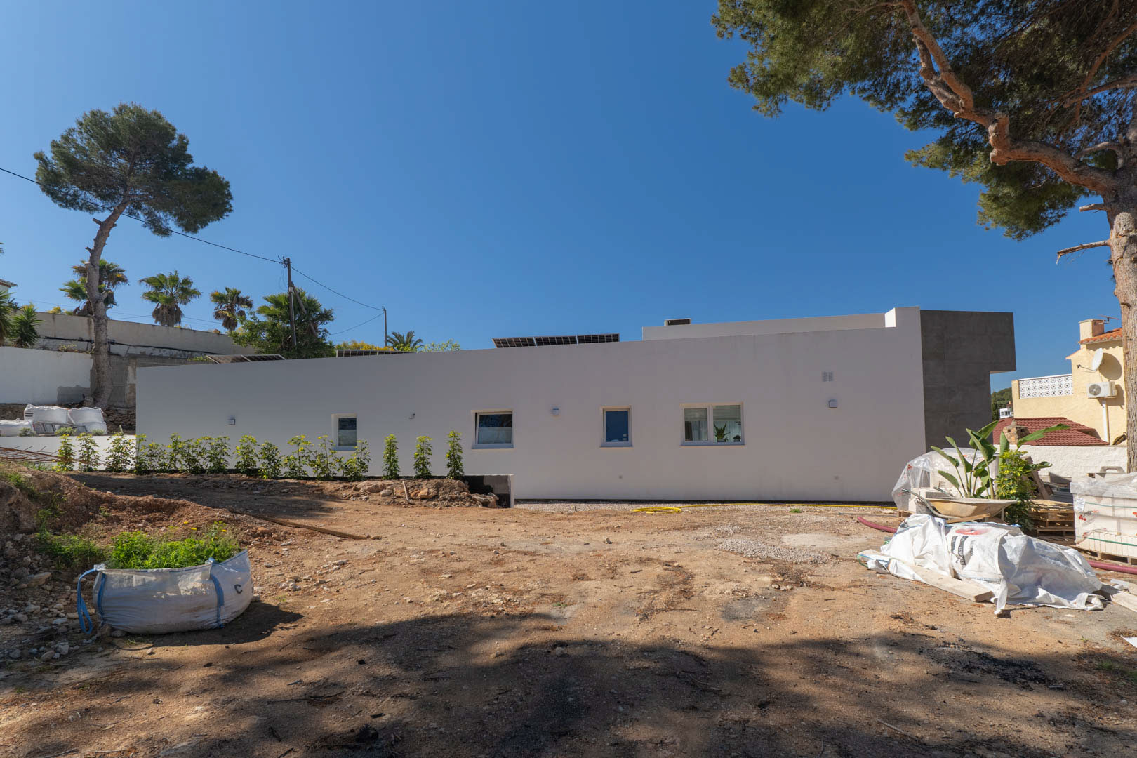 Façade latérale de la villa minimaliste avec panneaux solaires, illustrant le terrain extérieur en cours d'aménagement entouré de pins matures.