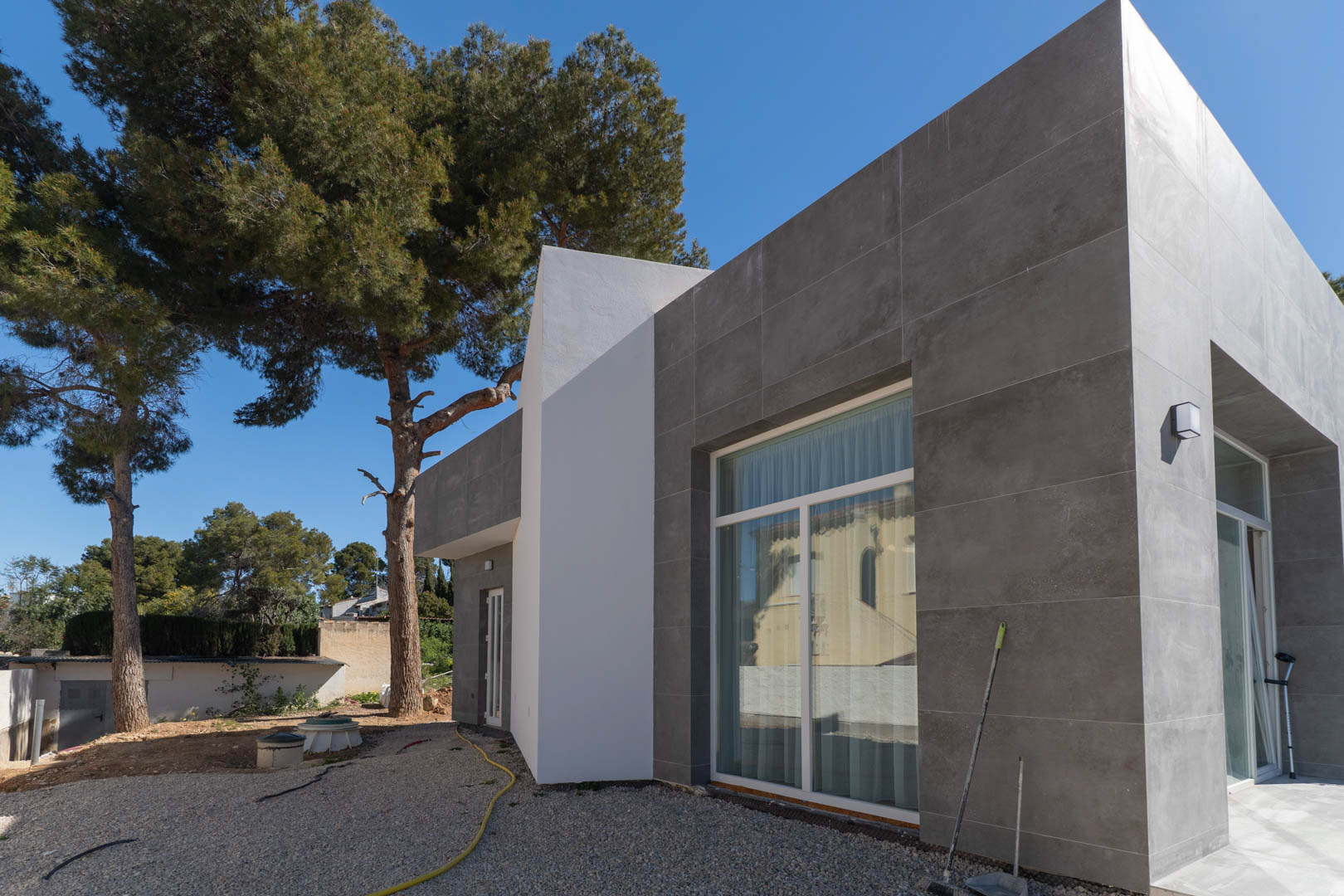 Façade minimaliste avec revêtement en céramique grise et grandes baies vitrées, entourée de pins matures et d'espaces extérieurs en cours de finition.