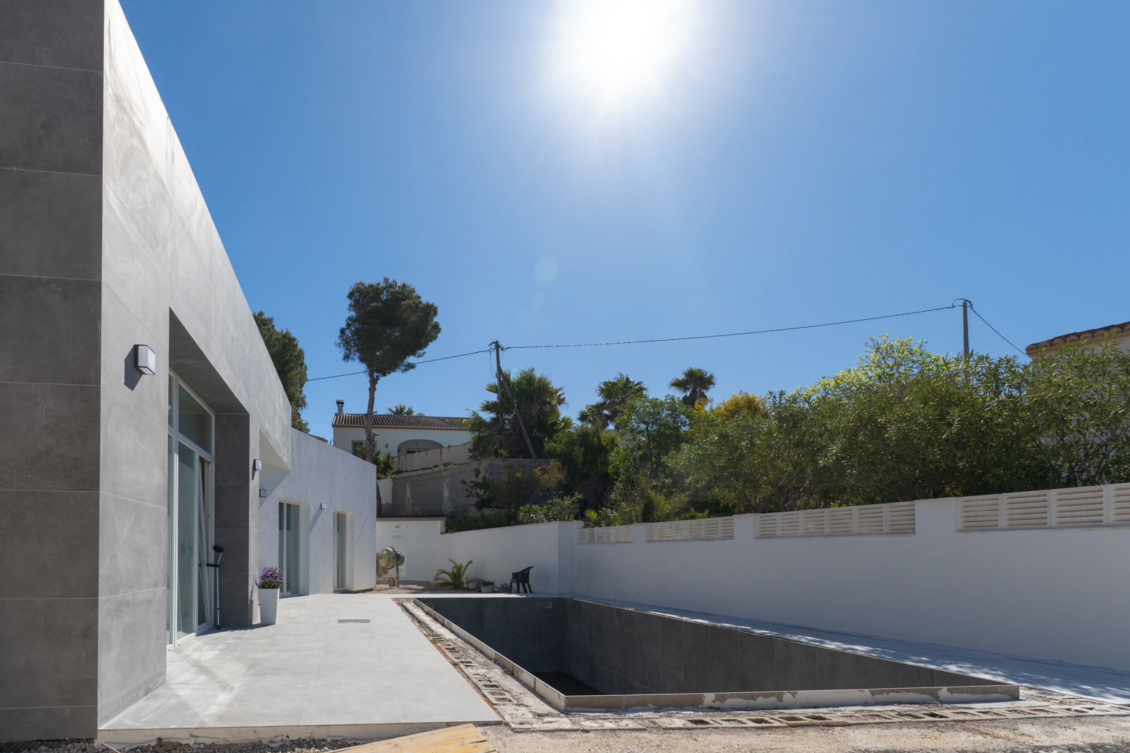 Façade minimaliste avec revêtement gris et zone de piscine en cours de finition, entourée d'un mur blanc sous un ensoleillement intense.