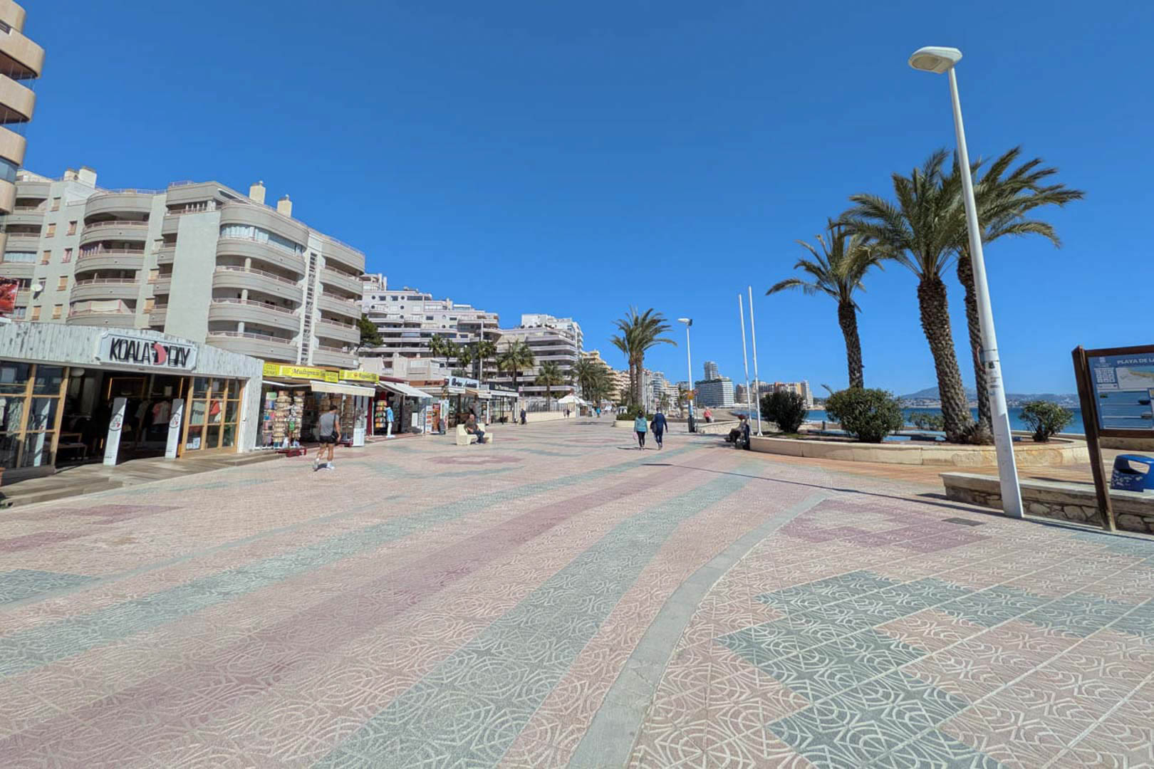Promenade maritime de Calpe avec pavage décoratif, bordée de boutiques, de palmiers et d'immeubles résidentiels sous un ciel azur.