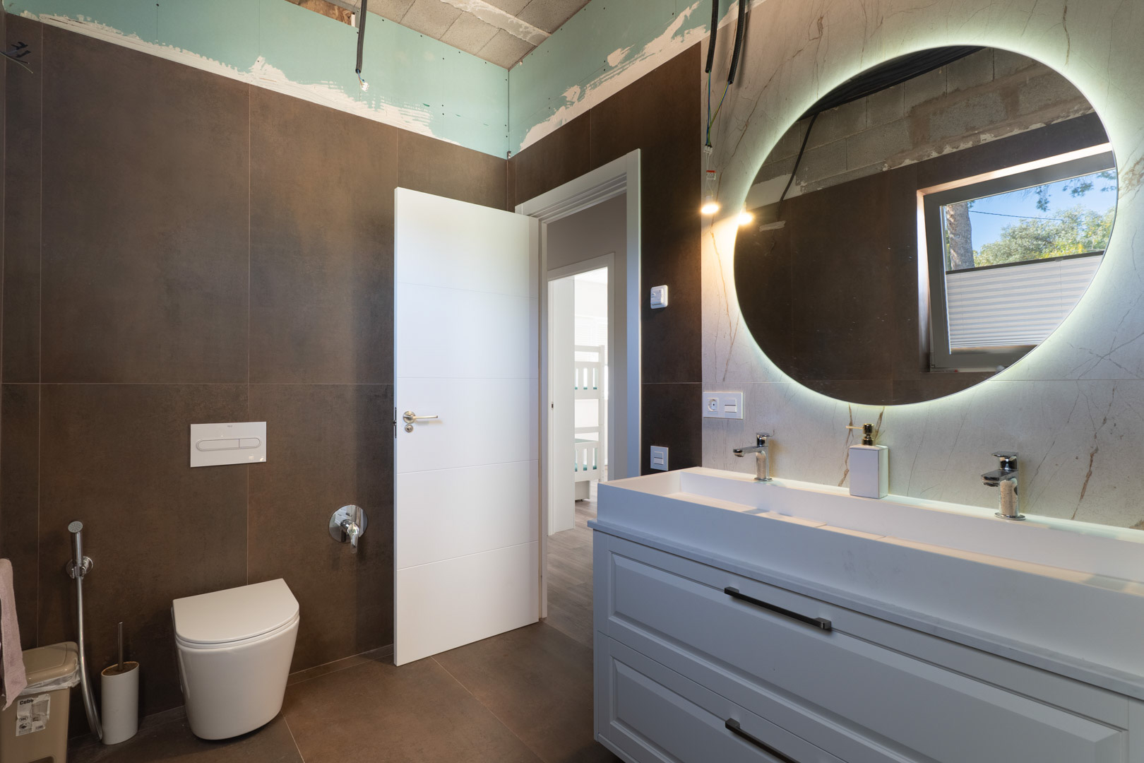Salle de bain contemporaine avec double vasque, miroir circulaire rétroéclairé et carrelage brun. Comprend un WC suspendu et des finitions de plafond en cours.